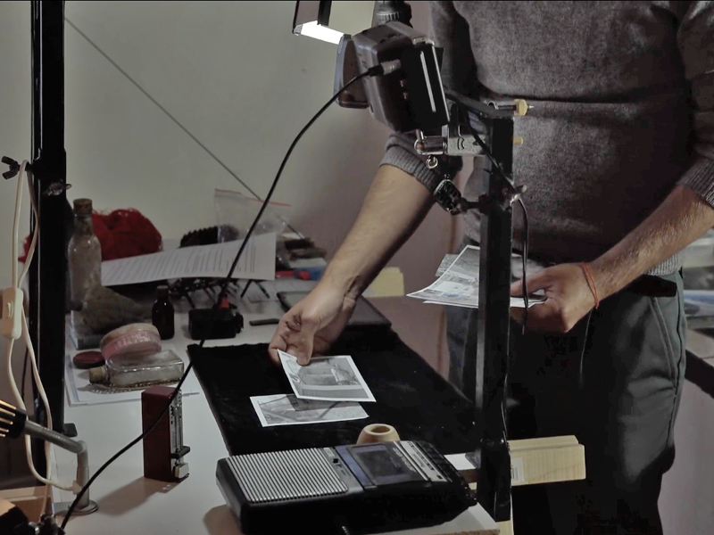 Eine Kamera und ein altes Aufnahmegerät auf einem Tisch. Eine Person legt gerade 2 Bilder auf einen schwarzen Untergrund.