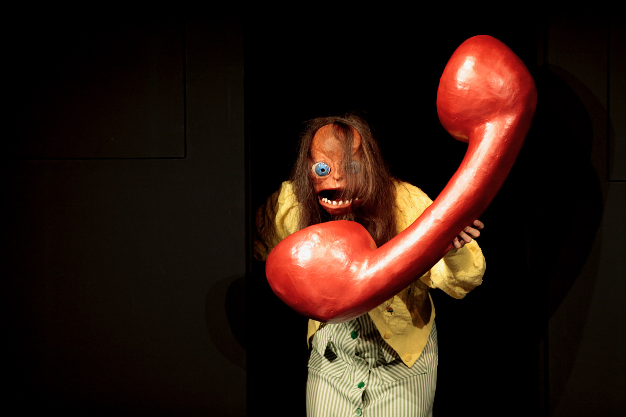 Eine Figur mit Maske hat einen übergroßen, roten Telefonhörer in der Hand.