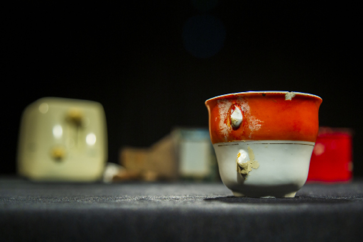 Verschiedene Gegenstände, darunter eine kaputte Teetasse und ein Toaster, stehen auf einer schwarzen Fläche. Die Tasse steht im Vordergrund und ist deutlich erkennbar, die anderen Objekte befinden sich verschwommen im Hintergrund.