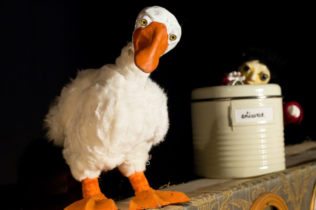 Figur einer Gans mit plüschig-weißem Gefieder und orangenem Schnabel steht seitlich vor einem grauweißen Topf mit Deckel. Der Deckel wird gerade von der Puppenfigur eines kleinen Jungen vielleicht angehoben, vielleicht  geschlossen.