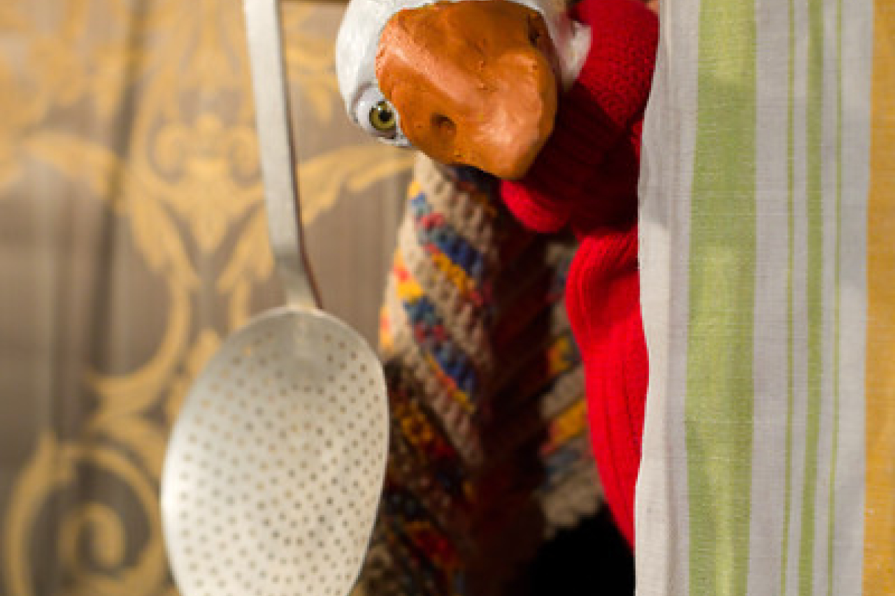 Nahaufnahme der Gans Auguste in rotem Pullover, neben ihr ein Schaumlöffel aus Aluminium. Rechts vorn im Bild gibt es ein Geschirrhandtuch mit hellgrünen Streifen.