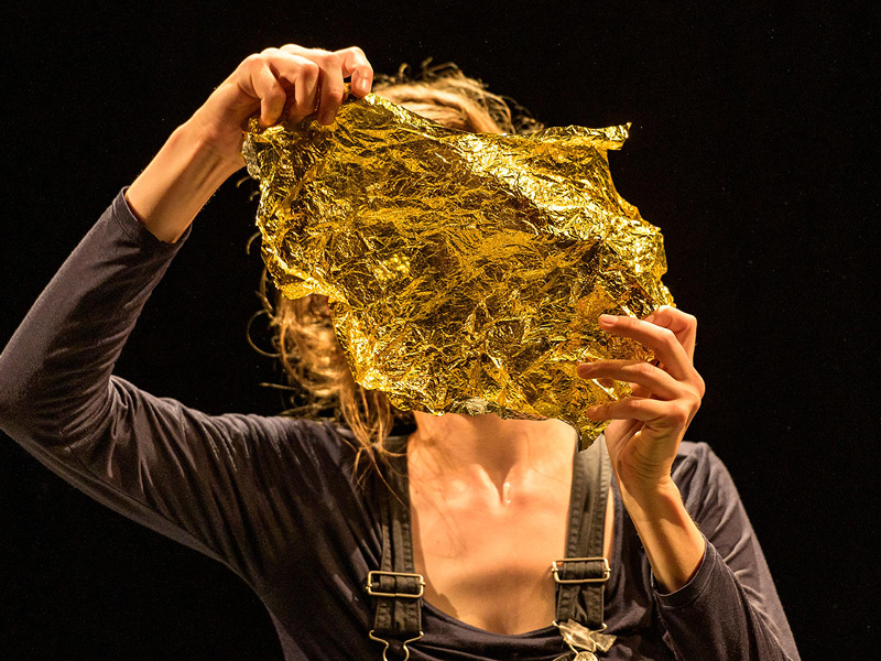 Eine dunkel gekleidete Frau mit Hosenträgern ist bis zum Rumpf abgebildet. Sie hält sich mit beiden Händen ein Stück goldene Rettungsfolie vor das Gesicht.