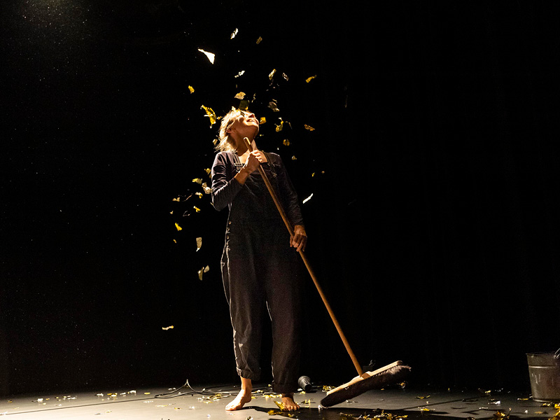 Performerin mit Besen in  der Hand auf dunkler Bühne. Über sie regnet es goldene Folienflocken. Am rechten Bildrand sieht man ein Stück von einem Eimer.