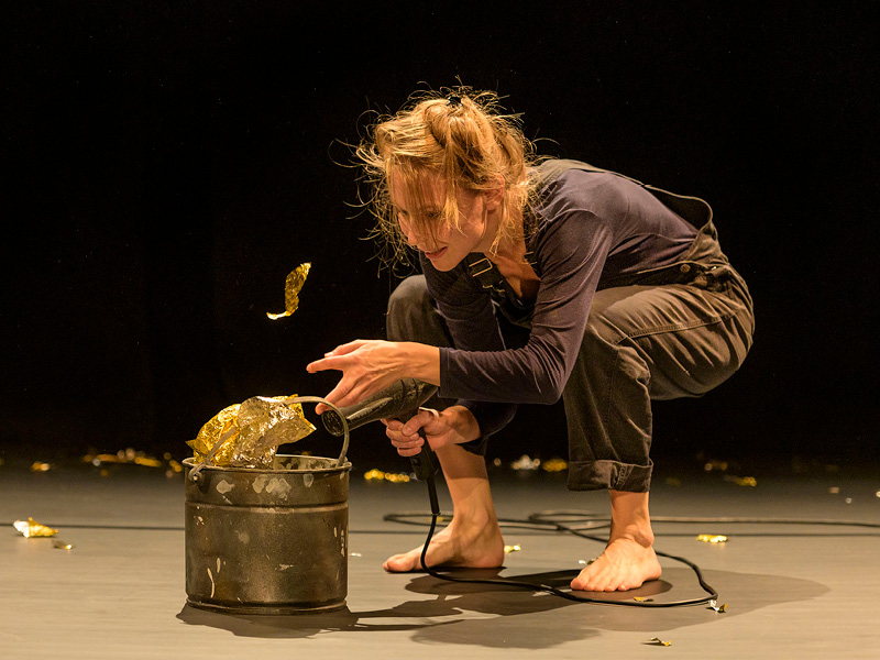 Eine Performerin mit Hose, blauem Sweatshirt und zum seitlichen Zopf gebundenen langen Haaren hockt barfuß vor einem Eimer, aus dem Glitzerfolie zu fliegen scheint. Im Hintergrund liegen weitere Fetzen von Glitzerfolie im Bühnendunkel.