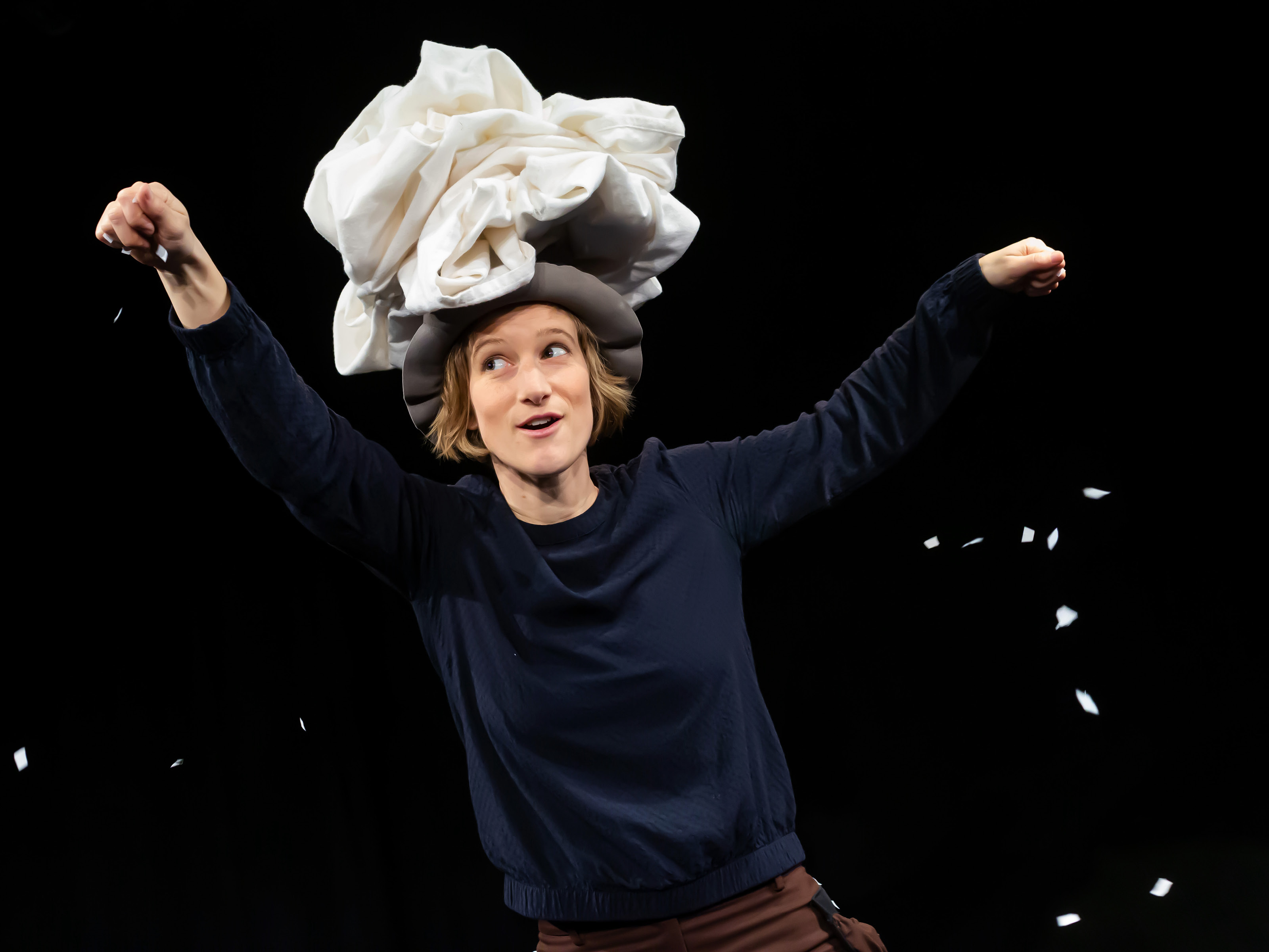 A woman in a white opulent cloth hat lets white scraps of paper trickle from her hands like snow onto the floor.