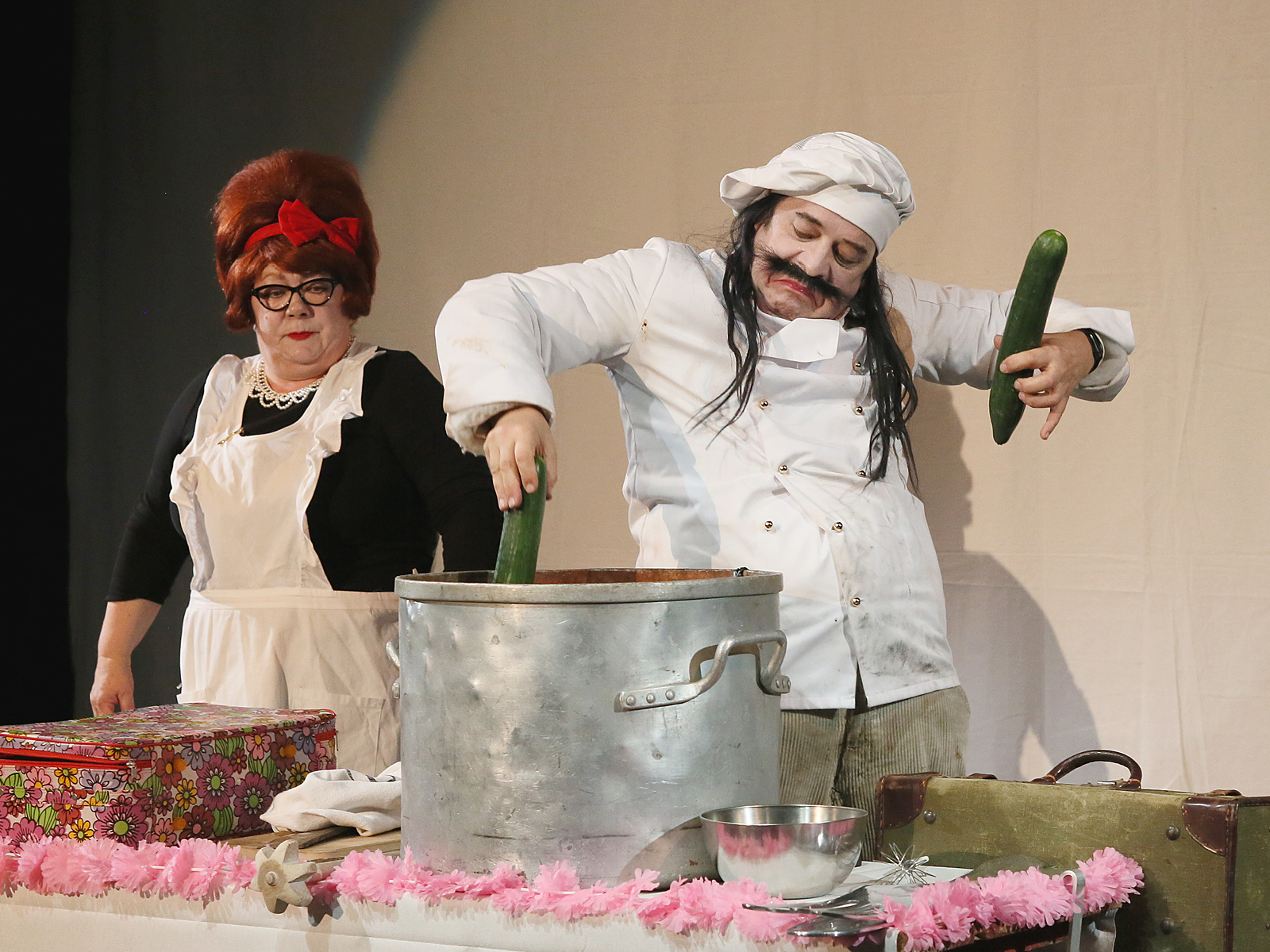 Langhaariger Koch mit Schnurrbart und Gurken in den Händen rührt damit in einem großen Alu-Topf. Frau mit Schürze steht daneben.