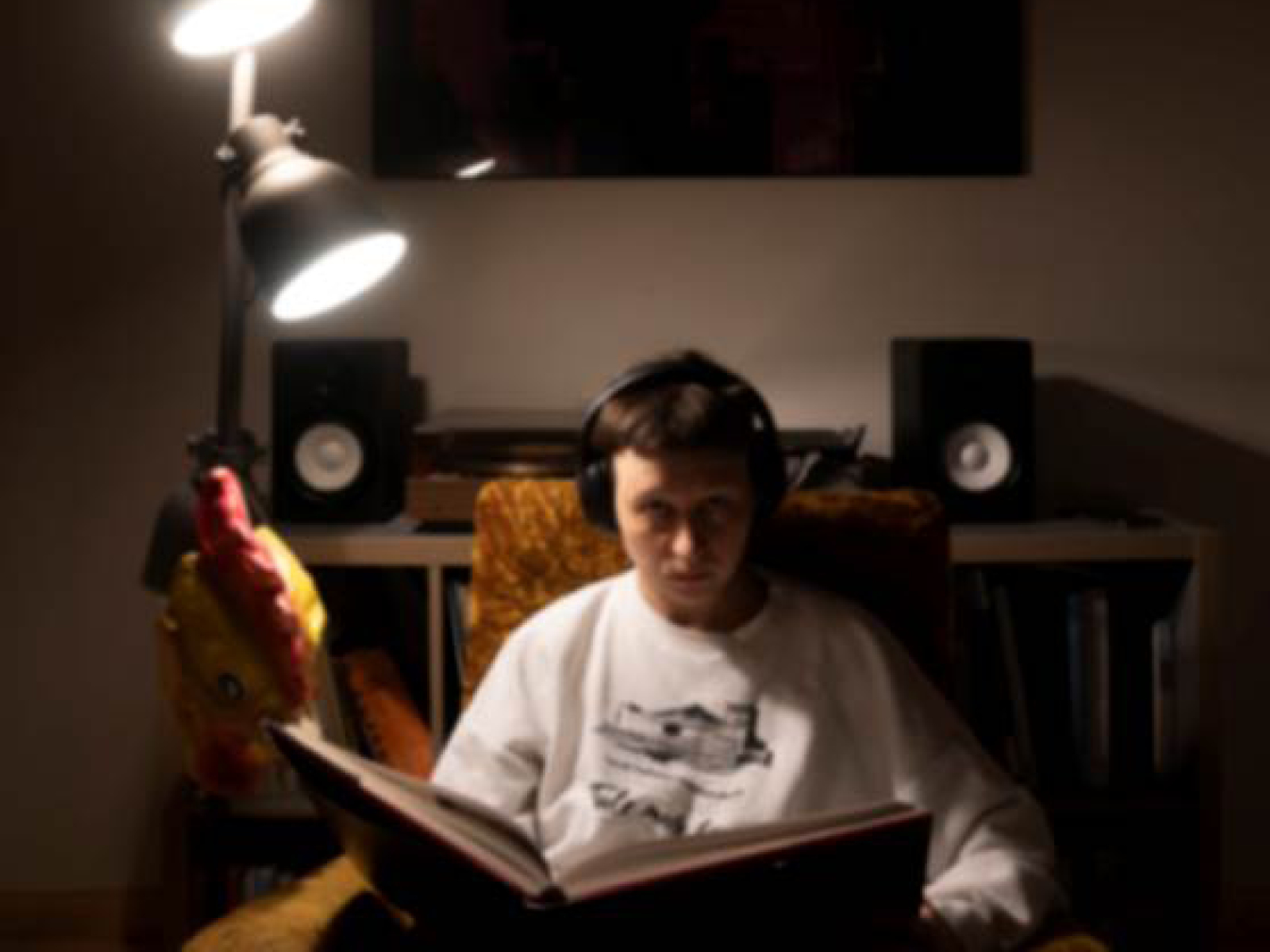 FRau in weißem Pulli und mit KOpfhörern schaut direkt in die Kamera. Auf ihrem Schoss ein aufgeschlagenes Buch. Im Setting eines normalen Wohnzimmers.