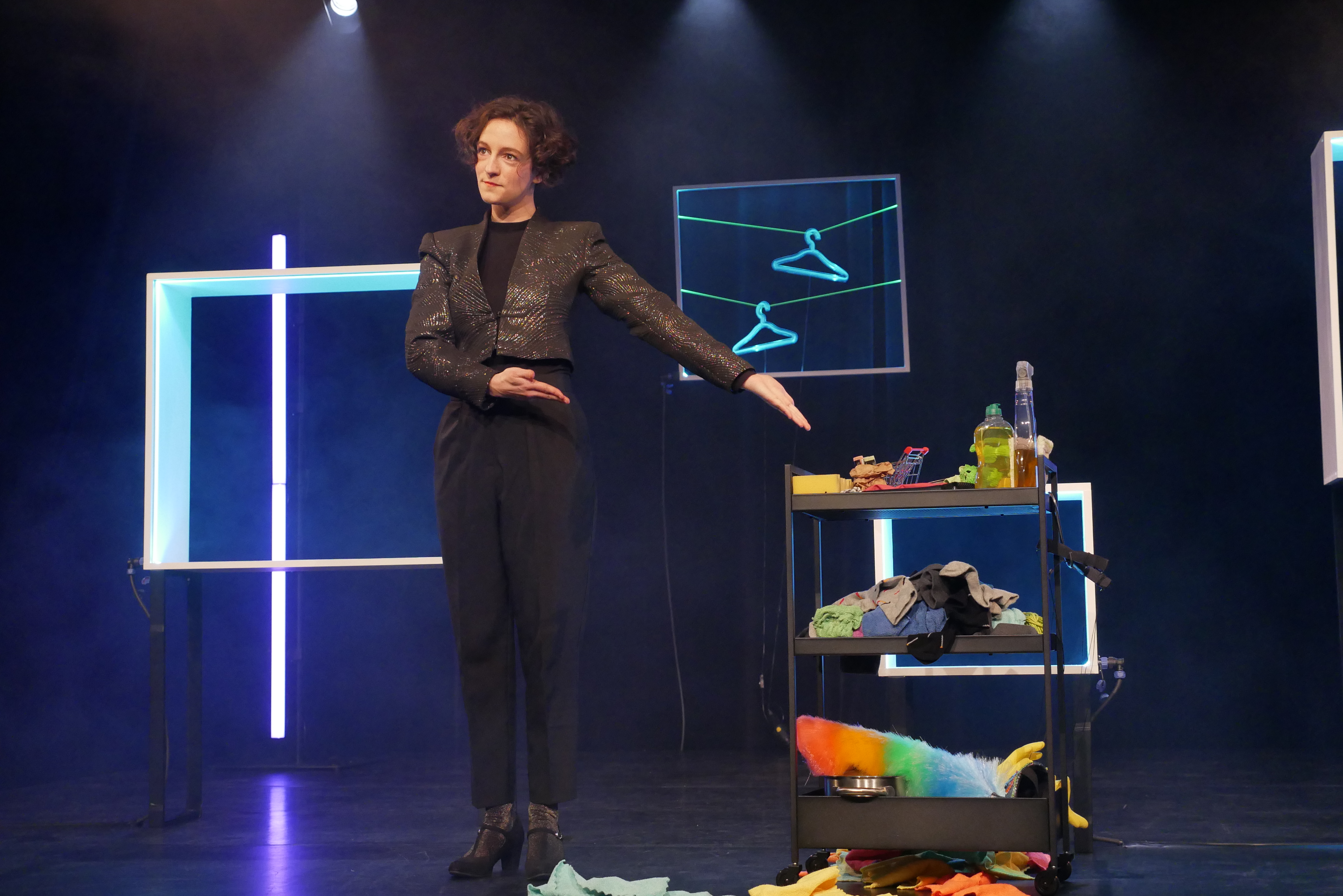 Josephine Hock, ganz in schwarz mit Glitzerjacke und hohen Schuhen, zeigt mit ihren Armen auf einen Putzwagen, der neben ihr steht und mit jeder Menge Putzutensilien bestückt ist. Im Hintergrund hängen als Deko Kleiderbügel.