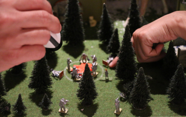 Zwei Hände interagieren mit einem kleinen Modell-Set in einem Tannenwäldchen mit einer Gruppe an menschlichen Figuren.