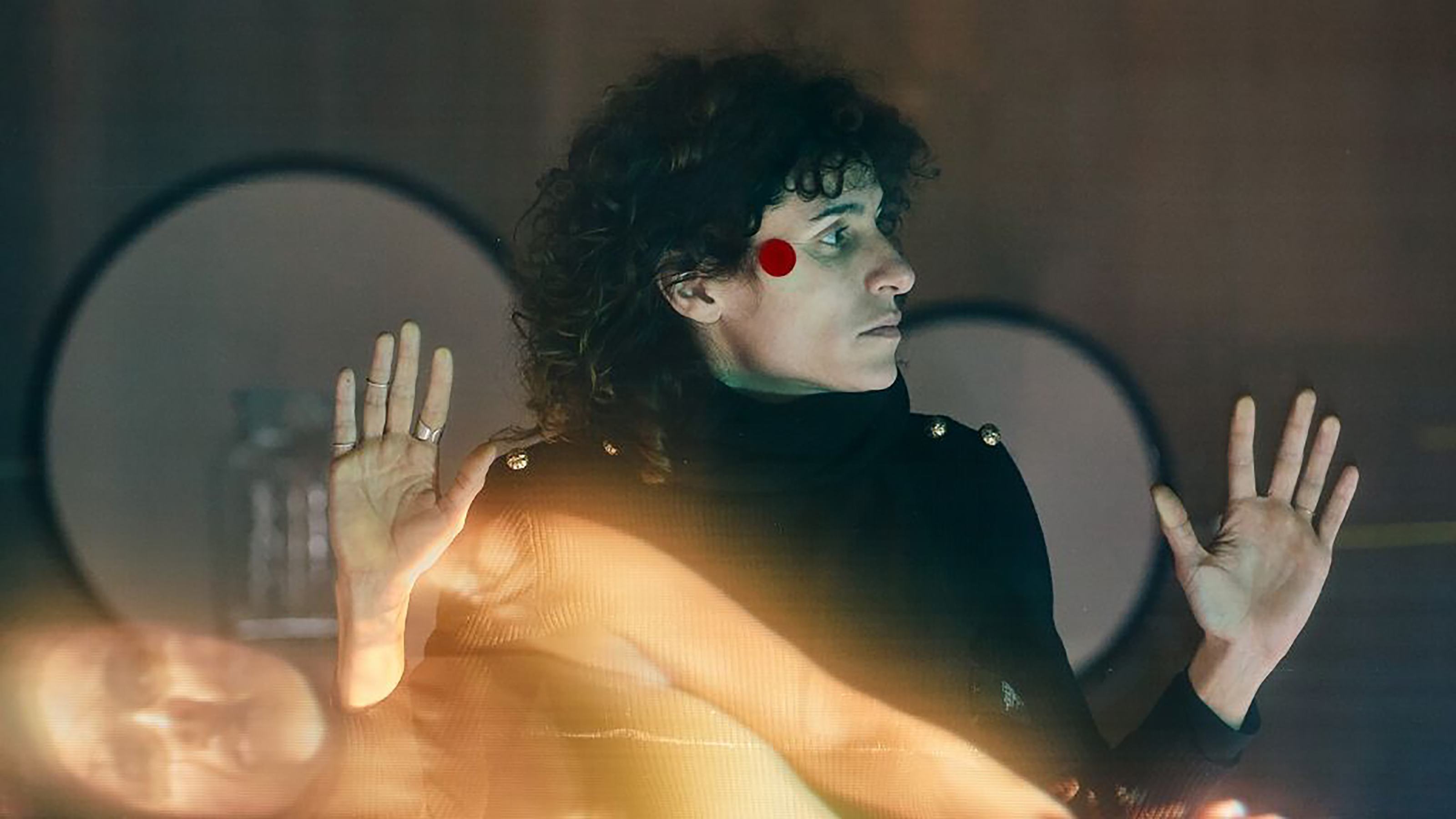 Eine Frau lehnt an einer Glasscheibe auf der die Projektion einer anderen Frau zu sehen ist. Hinter ihr stehen große Boxen.