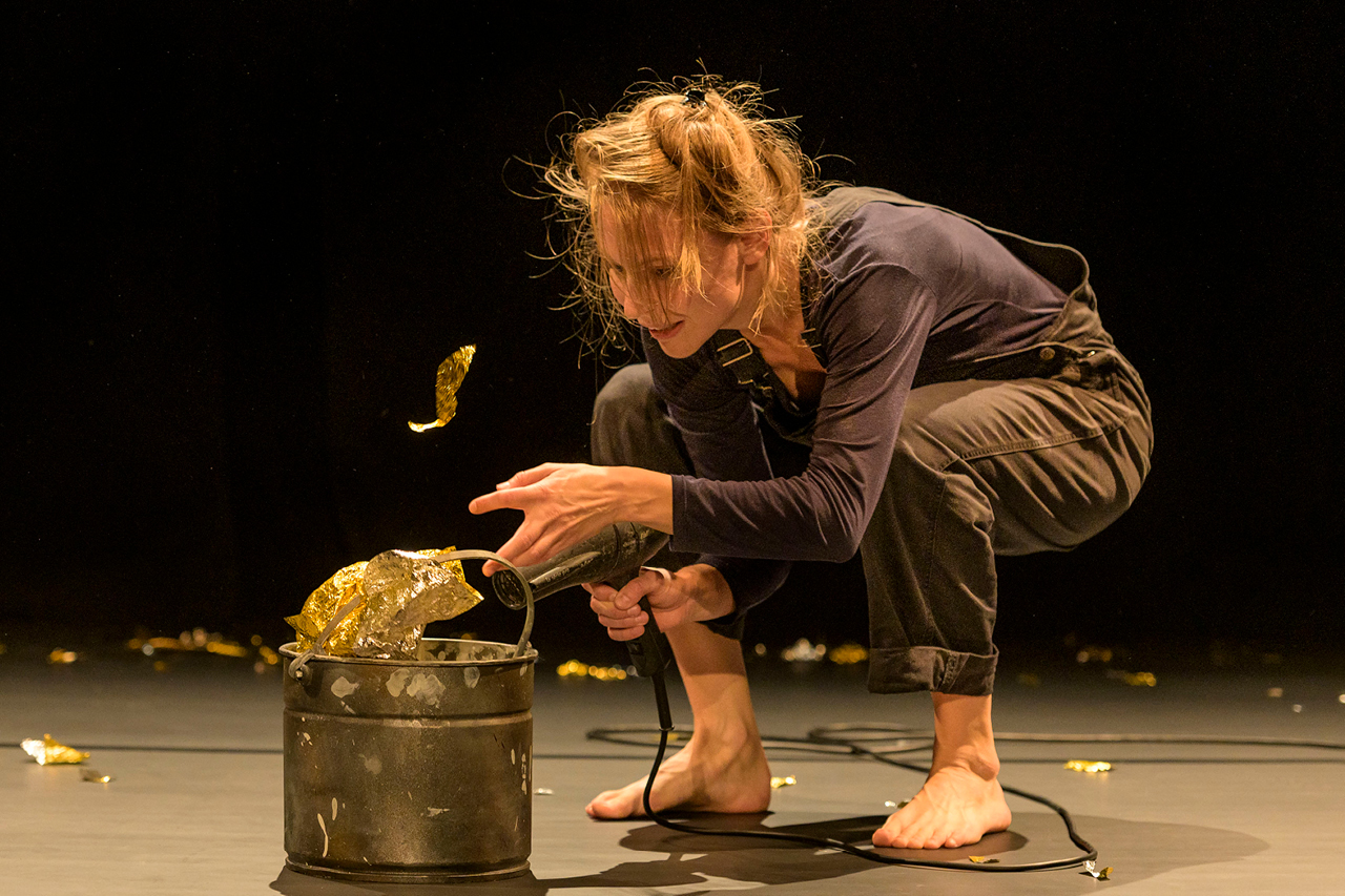 Eine Performerin mit Hose, blauem Sweatshirt und zum seitlichen Zopf gebundenen langen Haaren hockt barfuß vor einem Eimer, aus dem Glitzerfolie zu fliegen scheint. Im Hintergrund liegen weitere Fetzen von Glitzerfolie im Bühnendunkel.