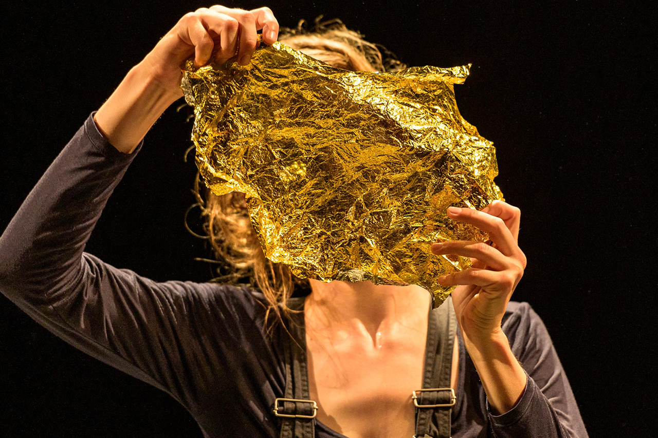 Eine dunkel gekleidete Frau mit Hosenträgern ist bis zum Rumpf abgebildet. Sie hält sich mit beiden Händen ein Stück goldene Rettungsfolie vor das Gesicht.