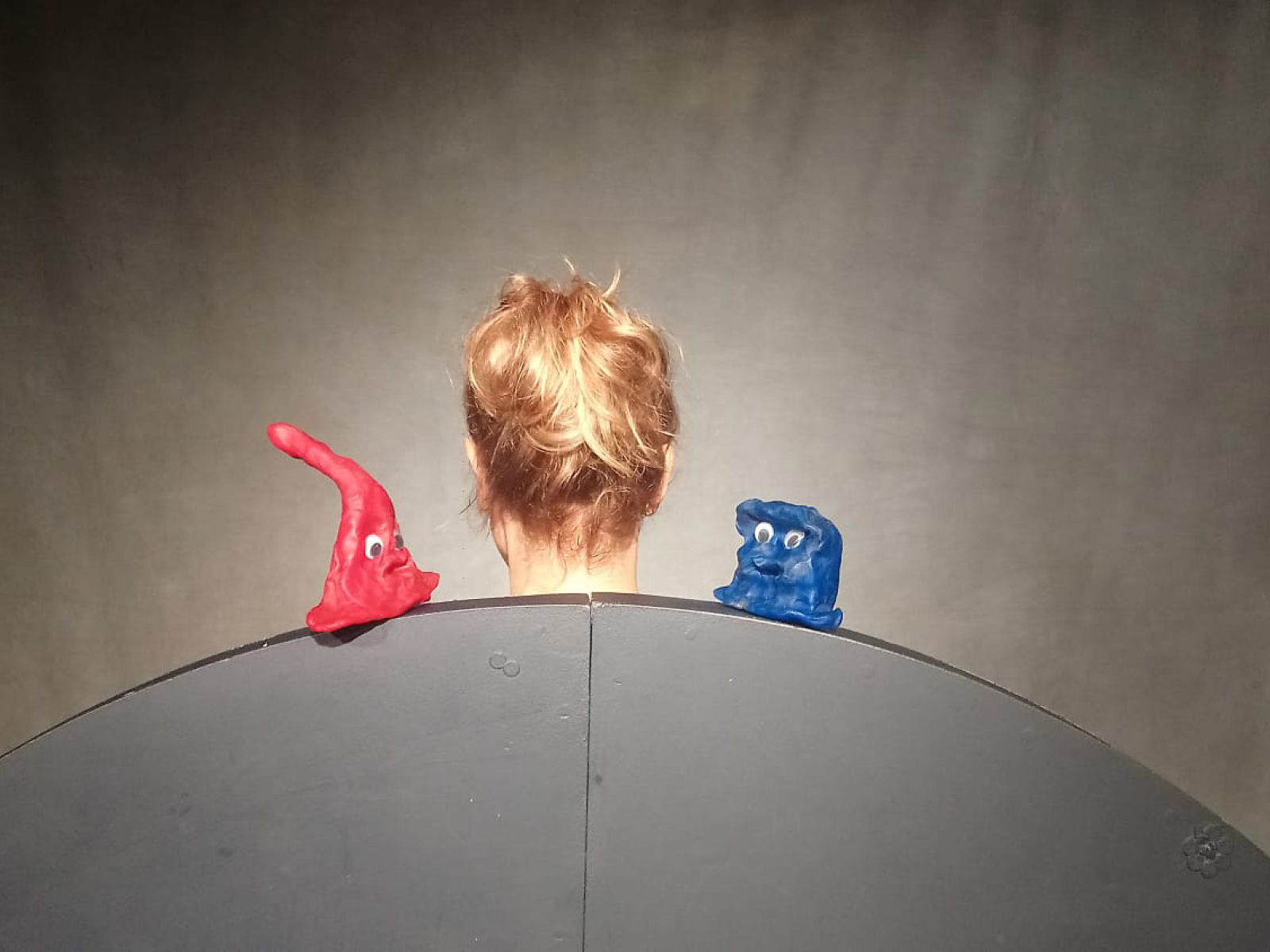 Ein runder Horizint. Darauf eine blaue und eine rote Knetfigur mit Augen. Dazwischen der Hinterkopf einer Spielerin.