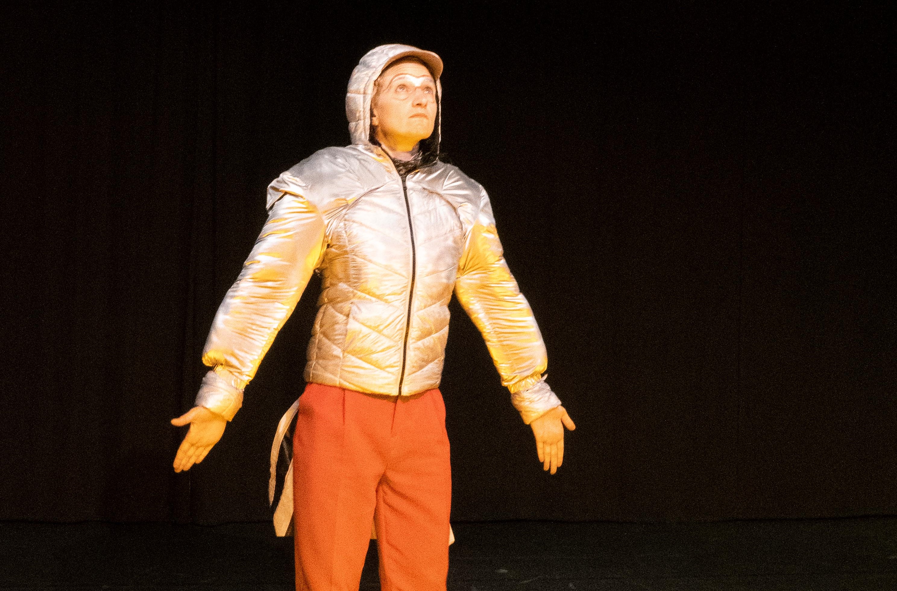 A person in a silver hooded jacket and aviator goggles has his arms spread out like wings for takeoff and is still looking up.