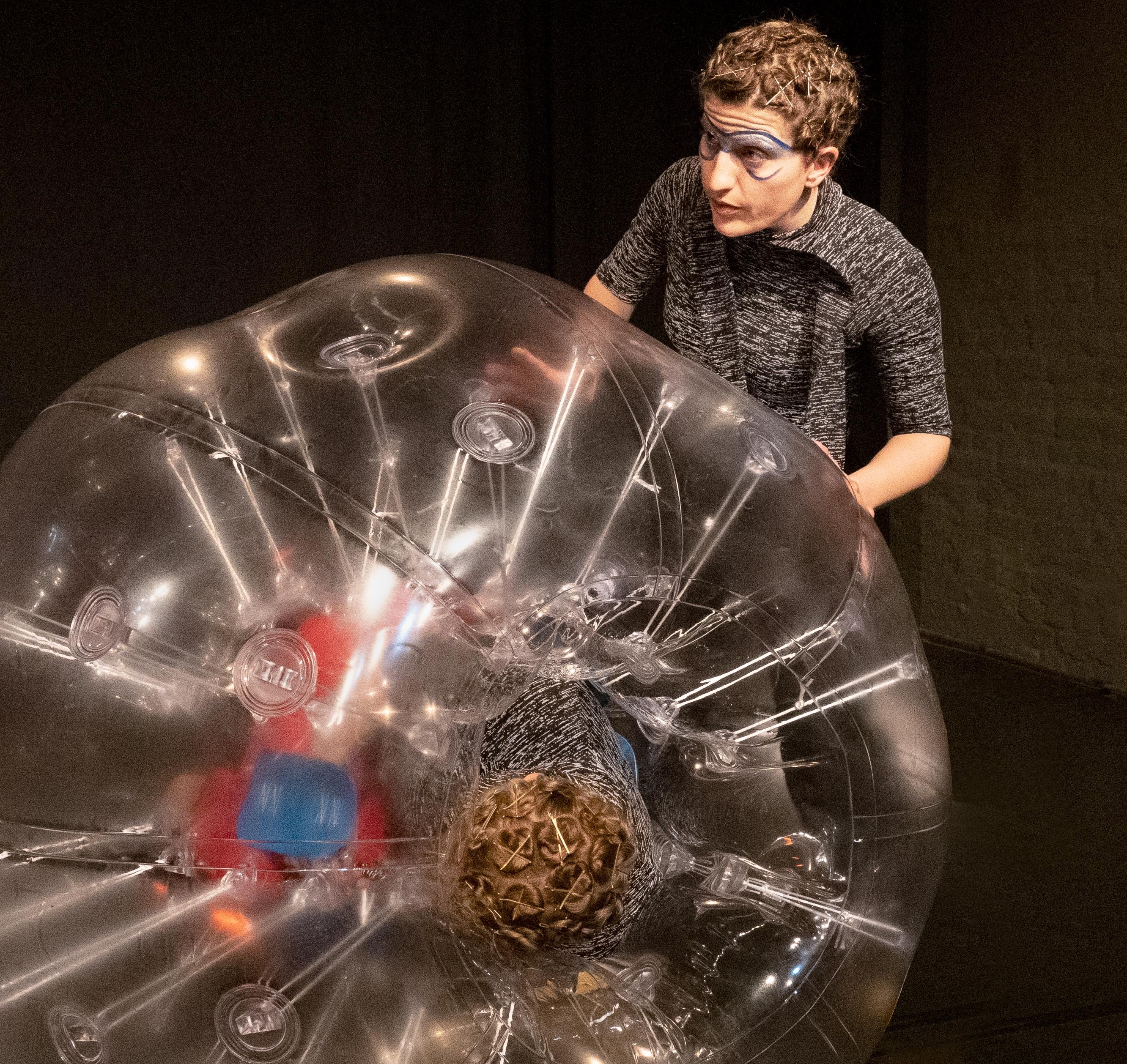 A person wearing painted-on aviator goggles rolls a person across the floor in a human-sized ball of air.