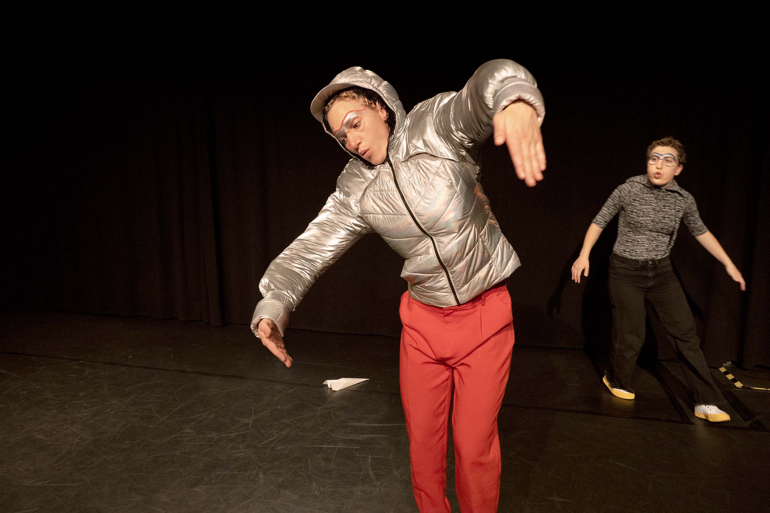 A person in a silver hooded jacket has his arms outstretched like wings and puts them with the upper body in the curve. Behind her is a person with a surprised expression.