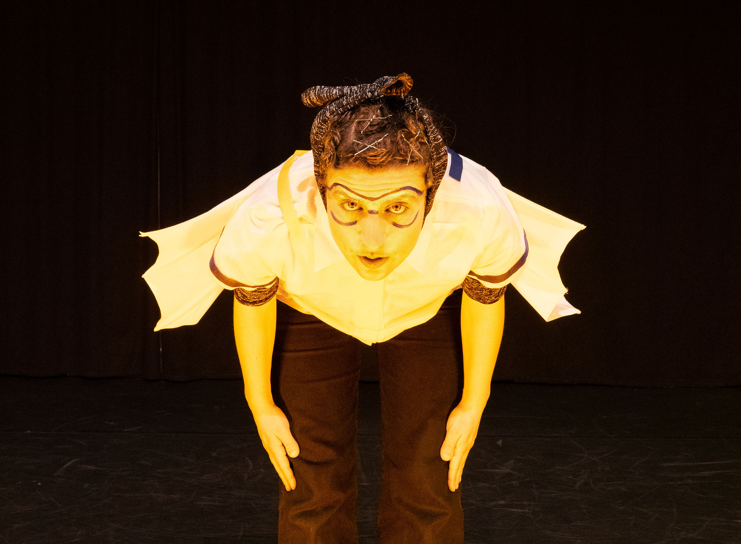 A yellow lit man with painted on aviator goggles has his upper body bent as if for flight takeoff. On his costume are wings.