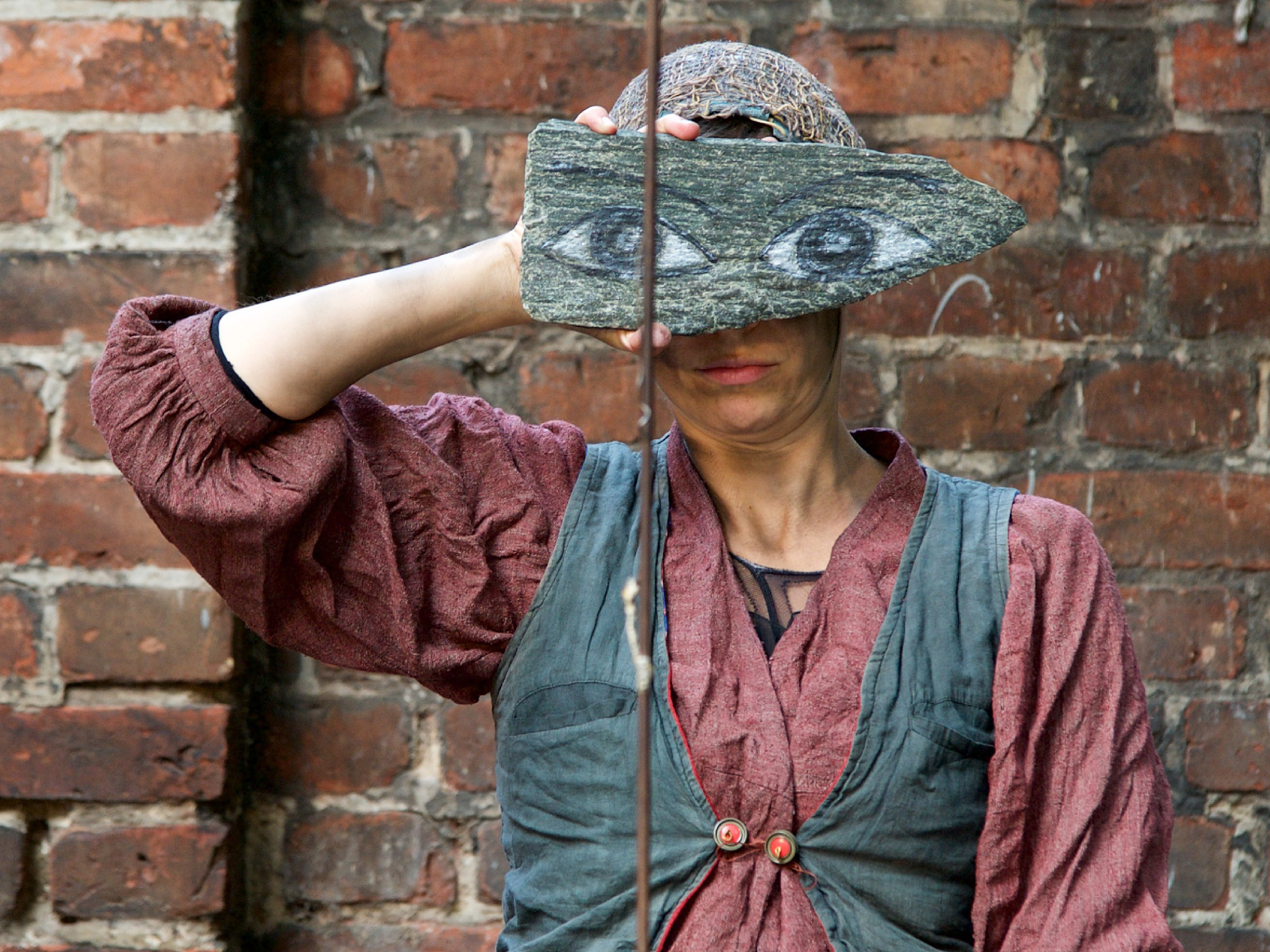 A woman with an opulent red linen blouse and a blue vest over it holds a piece of wood on which two eyes are painted in front of her face. You can see only her mouth and the base of a headscarf.