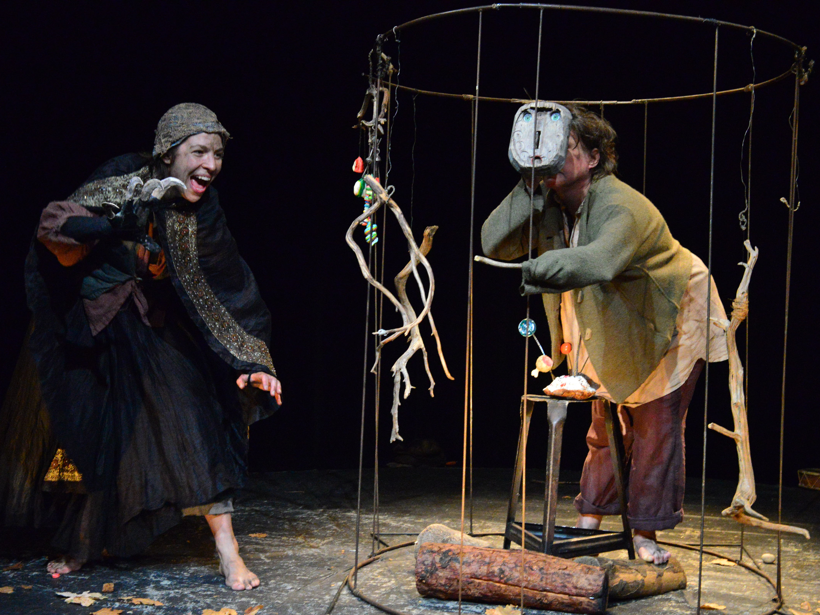 Hansel is in a round cage made of metal bars. He holds one of the wooden masks painted with a face in front of his face. He stretches his supposed finger through the bars. However, it is a thin branch. The witch is barefoot on her way to him.