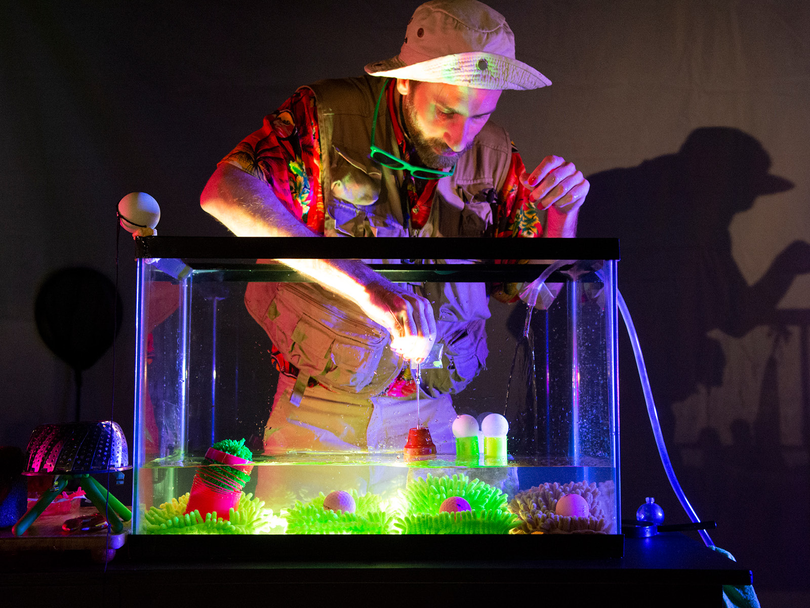 Robin Leveroos drückt eine Pipette voll Wasser in ein bunt beleuchtetes Aquarium vor ihm, das mit Gummiobjekten gefüllt ist, die wie abstrahierte Austern aussehen und in sich rosafarbene Gummibälle tragen. Die Szene erinnert an eine Unterwasserlandschaft mit Korallen und Muscheln.