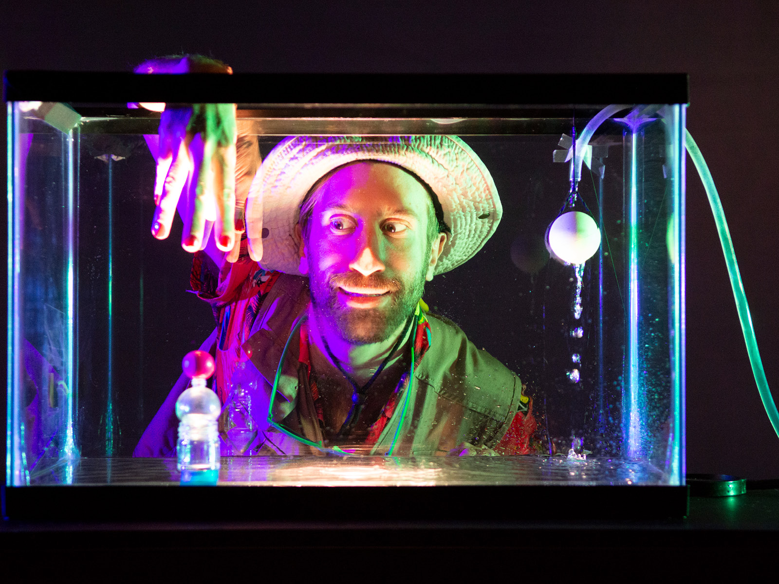 Hinter einem hell Magenta und Grün beleuchteten Aquarium sitzt Robin Leveroos und schaut lächelnd auf eine abstrakte Figur innerhalb des Aquariums. Robins Hand streckt sich von oben nach der Figur aus. Das Aquarium wird gerade mit einem kleinen Waserschlauch aufgefüllt und die ersten Tropfen fallen auf den Boden des Aquariums.