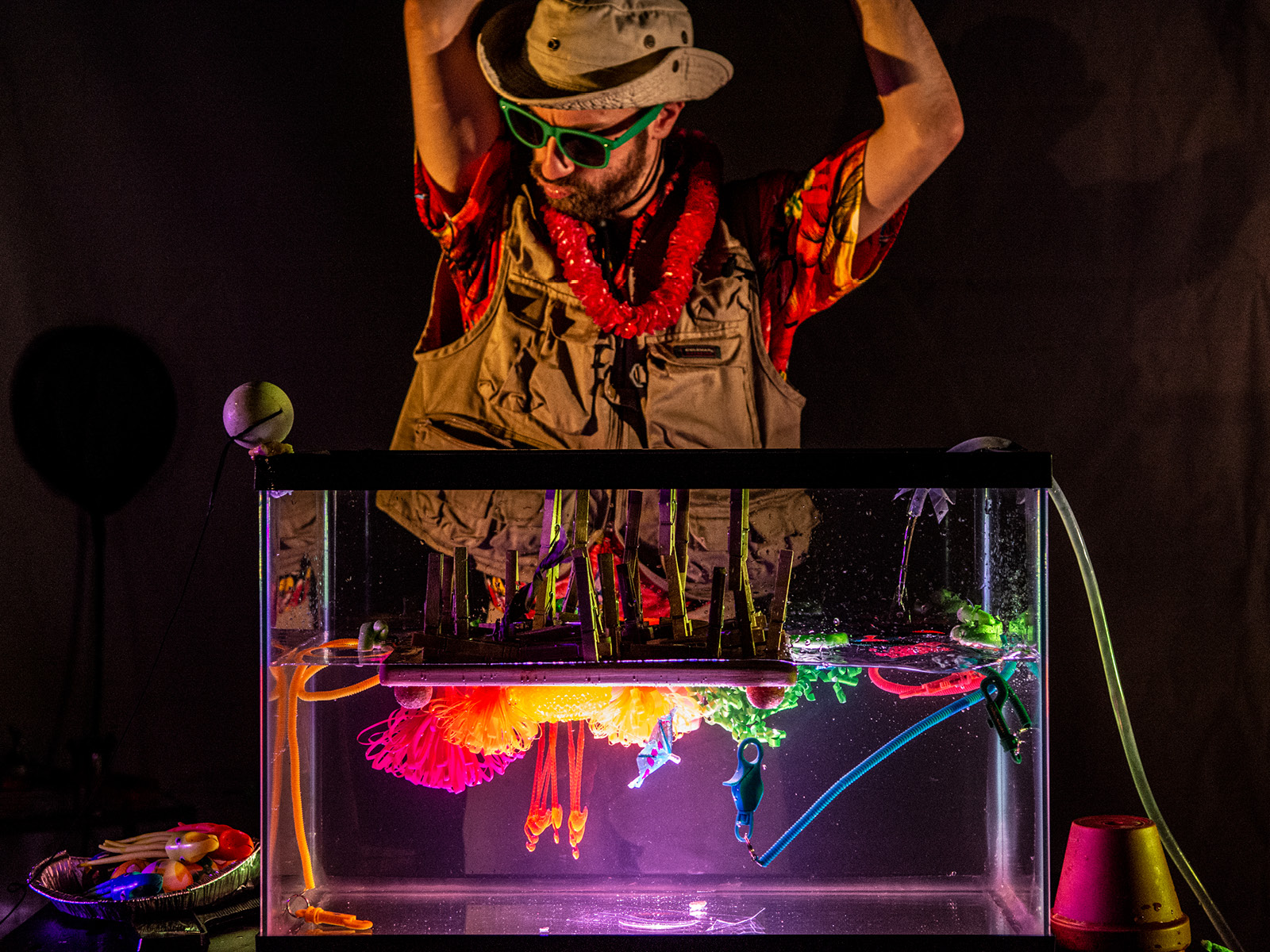 Robin Leveroos steht mit erhobenen Händen vor einem erleuchteten Auqarium, das mit bunten Dingen verschiedenster Farben und Konsistenten gefüllt ist. Robin trägt eine Sonnenbrille, einen Blumenkranz um den Hals, eine Anglerweste, einen Hut und ein Hawaiihemd.