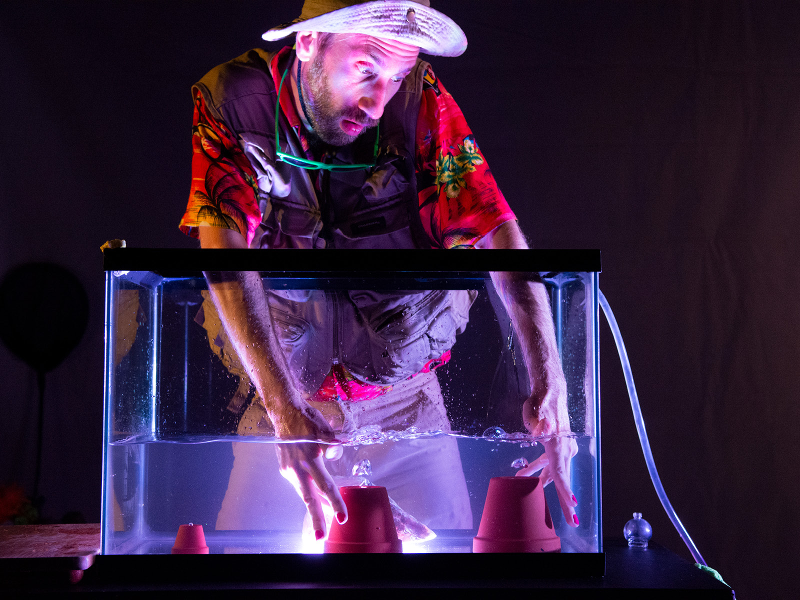 In einem hell beleuchteten Aquarium mit zwei umgekehrten Blumentöpfen stecken die Hände von Robin Leveroos und wühlen die Wasseroberfläche auf. Robin trägt ein stereotypes Entdeckeroutfit, bestehend aus Kakihosen, eine Weste mit vielen Taschen, einen Hut mit großer Krempe und ein Hawaiihemd.