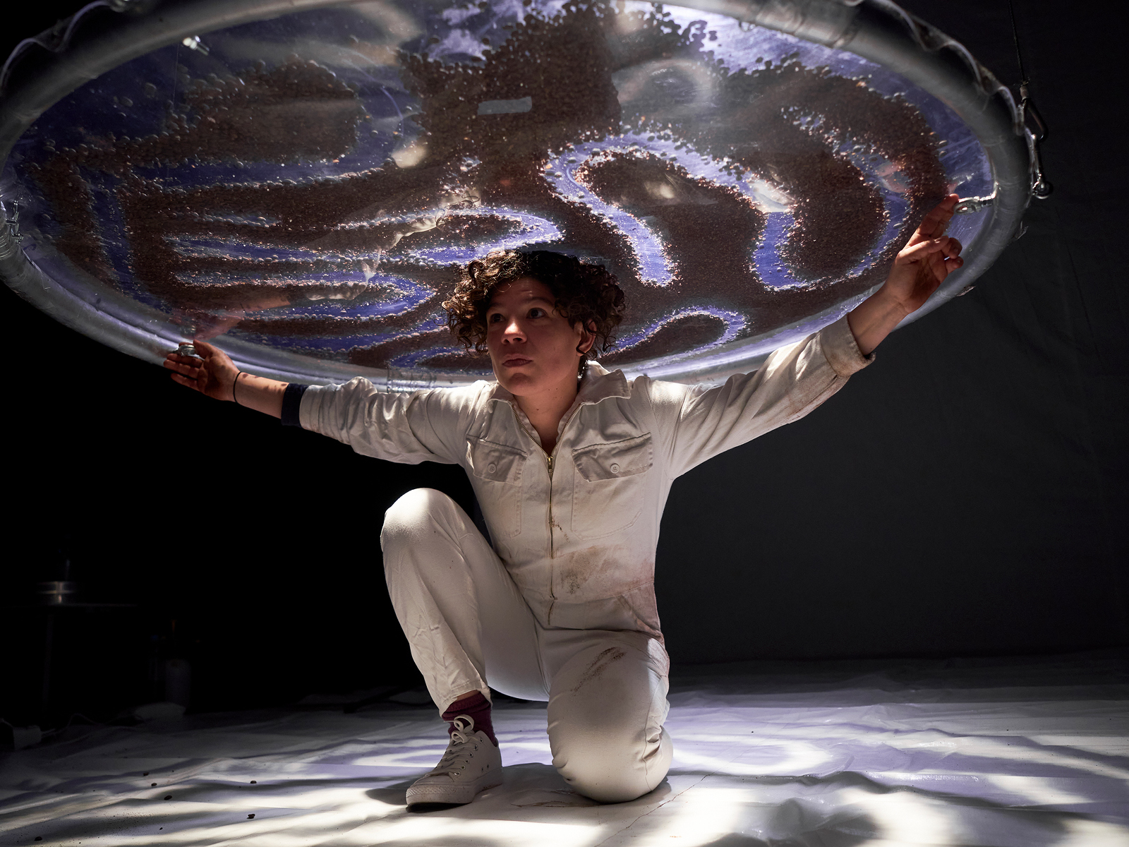 Unter einer blau beleuchteten Scheibenkonstruktion, die an Drähten von der Decke hängt, ist die Performerin in weißem Ganzkörperanzug abgebildet. Sie kniet. Auf der Scheibe sind Kaffeebohnen zu einem Muster zusammengeschoben.