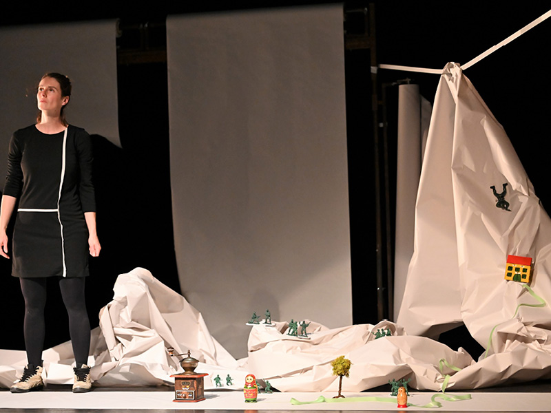 Eine schwarz gekleidete Frau steht vor einer Berglandschaft aus Segeltuch mit Miniaturfiguren. Neben ihr steht eine Kaffeemühle und eine Babuschka.