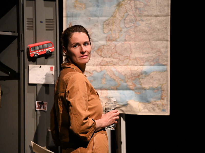 Eine mittelalte Frau in einem beigen Overall schaut über die Schulter an der Kamera vorbei. Hinter ihr ist eine Landkarte von Europa angebracht.