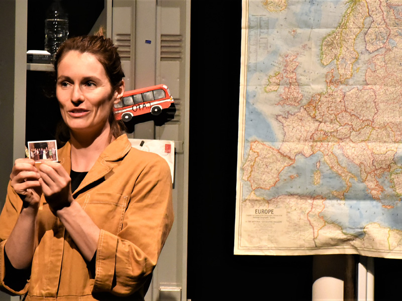 Eine mittelalte Frau in einem beigen Overall hält ein Gruppenfoto in der Hand und schat zu den Zuschauern. Sie steht vor einer Europakarte, neben der ein Omnibusmagnet hängt.