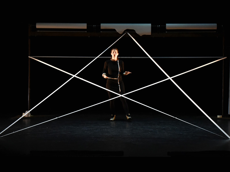 Eine schwarz gekleidete Frau steht auf einer dunklen Bühne und ist von einem Pentagram aus Schnüren umgeben.