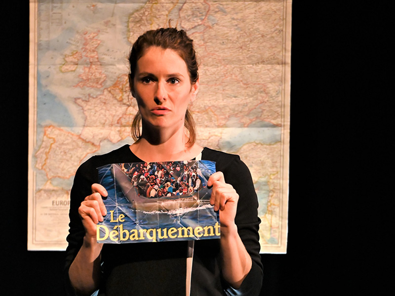 Die Frau hält mit traurigem Gesicht einen Magazinausschnitt mit dem Bild eines Flüchlingsbootes voller Menschen hoch, hinter ihr ist eine Landkarte von Europa.