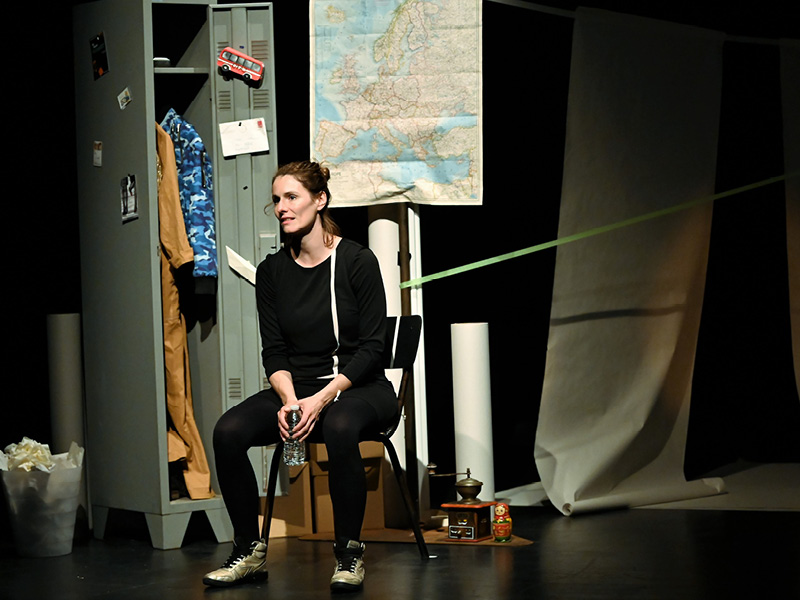 Eine mittelalte Frau sitzt umgeben von Requisiten neben einem Spind mit Klamotten. Hinter ihr ist eine Landkarte von Europa.