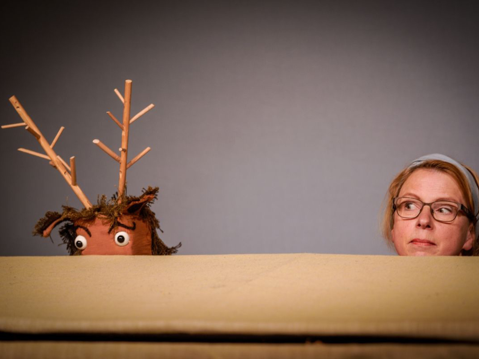 Über den Rand einer beige farbenen Fläche schaut ein Hirsch mit großen Augen, markanten, geschwungenen Augenbrauchen, einem Geweih aus kleinen Ästen und wuscheligen Ohren. Neben ihm lugt die Puppenspielerin Susanne Olbrich über den Rand.