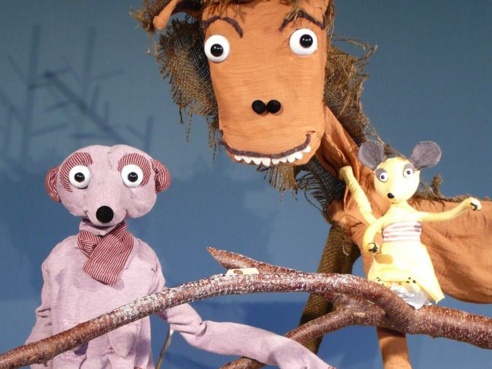 Die Figuren der HauptprotagonistInnen der Inszenierung sind einem Familienfoto ähnlich im Bild platziert und schauen in die Kamera. Der Hirsch mit den markanten Augenbrauchen, dem zotteligen Fell und dem Überbiss steht hinten. Die Maus im zarten Kleidchen mit erhobenen Armen steht rechts auf einem Ast. Und das Erdmännchen mit den langen Armen steht links.