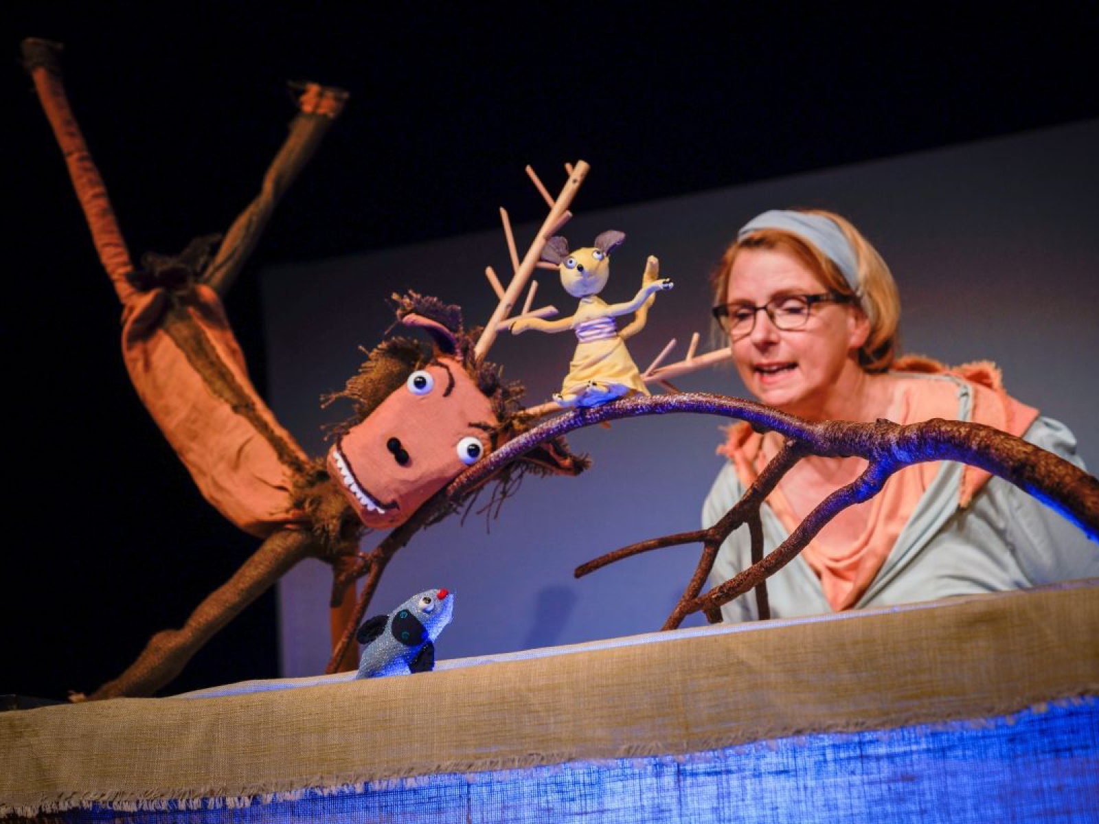 Der Hirsch hat seine Hinterbeine in die Luft geworfen und lächelt mit leichtem Überbiss in die Kamera. Mit seinem Kopf drückt er einen Ast hinterunter, auf dem eine Maus mit Kleidchen sitzt. Unterhalb des Astes schaut ein Fisch zu den Beiden herauf. Das Wasser seines Teiches beleuchtet den Ast, auf dem die Maus sitzt. Susanne Olbrich steht im Hintergrund und sagt etwas.