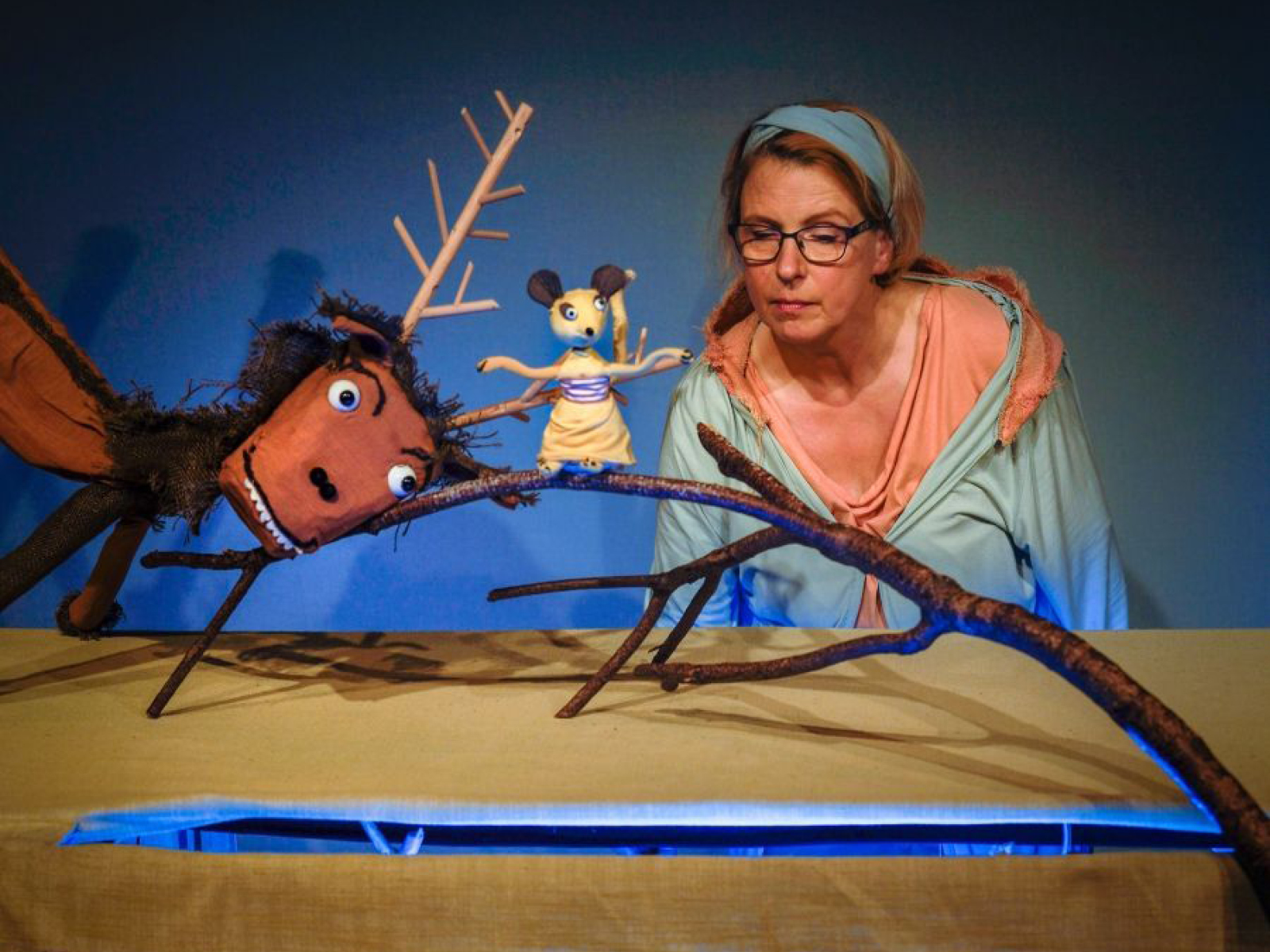 Der strubbelige Hirsch mit vereinzelten zotteligen Fellstücken, holzigem Geweih, markanten Augenbrauchen und leichtem Überbiss drückt mit seinem Kopf einen Ast hinunter, auf dem eine kleine Maus mit Kleidchen in sanften Beigefarben sitzt. Unter ihr ist ein Teich, aus dem ein Fisch hinauf schaut. Puppenspielerin Susanne Olbrich betrachtet die Szene aus dem Hintergrund.