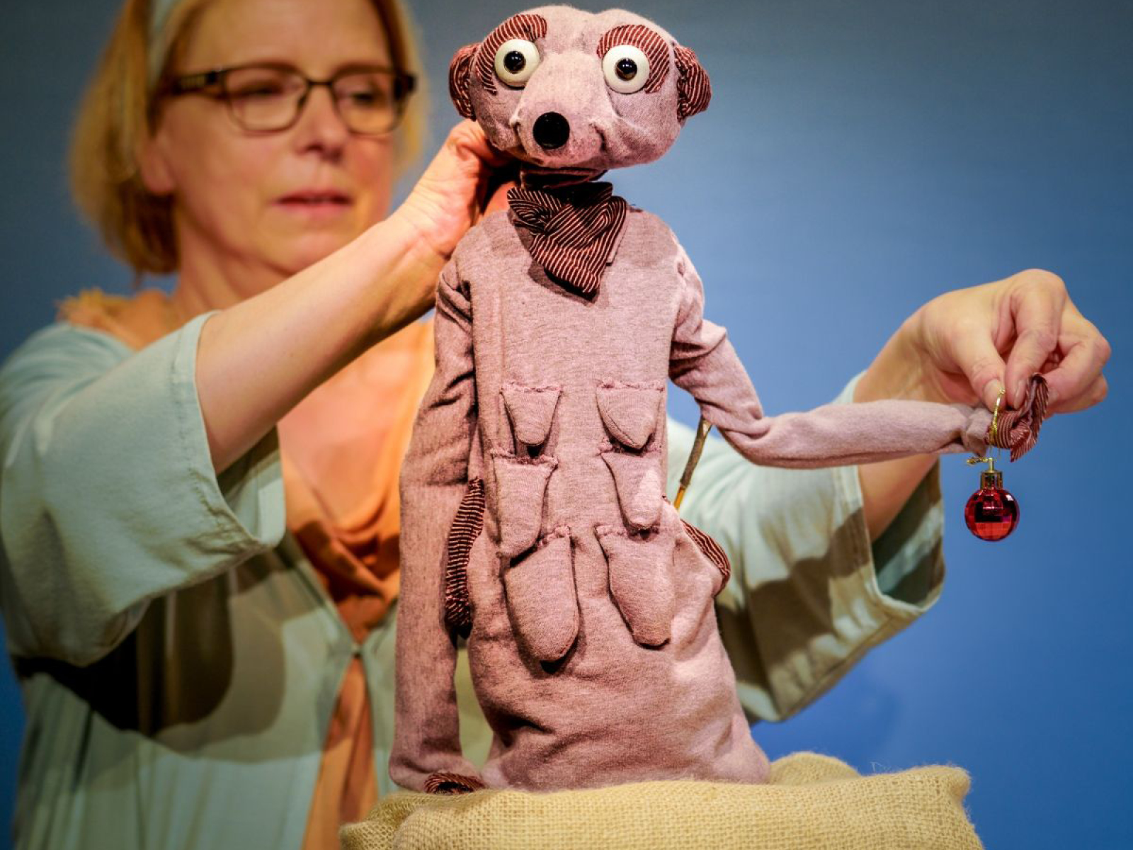 Ein Erdmännchen aus schlapprigem, rosafarbenem Stoff, mit ausgerpägten Zitzen und sehr langen Armen hält in der rechten Hand eine rote, glänzende Weihnachskugel. Die großen, runden Augen sind schauen neugierig richtig Kamera. Susanne Olbrich steht im Hintergrund und bewegt die Gliedmaßen des Erdmännchens.