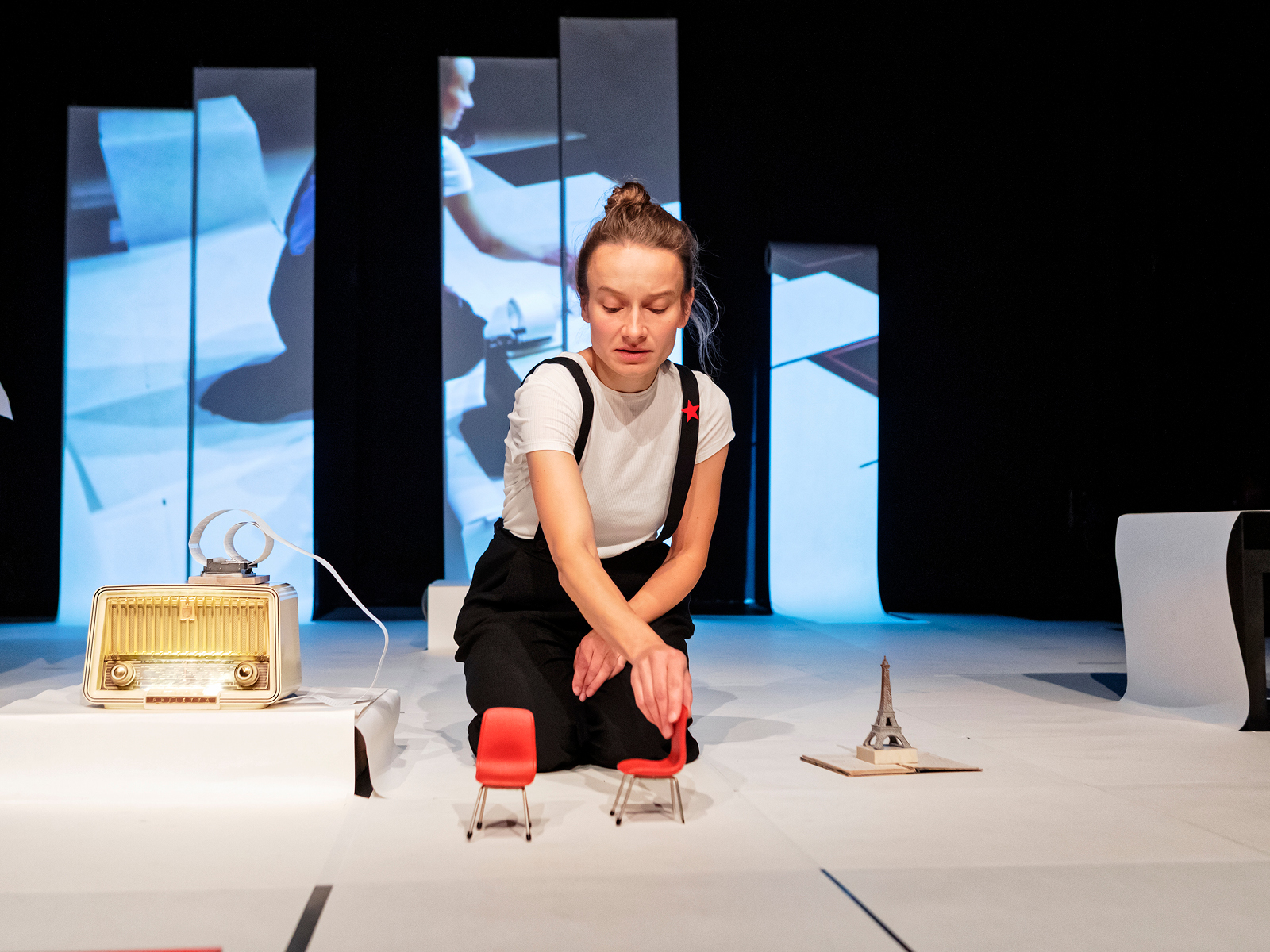 Spielerin auf der Bühne. Sie rückt gerade ein kleines rotes Modell-Stühlchen auf dem Boden zurecht, daneben befindet sich ein zweites sowe ein Miniatur-Eiffelturm. Links davon ein alter Radioemfänger, im Hintergrund eine Videoprojektion.