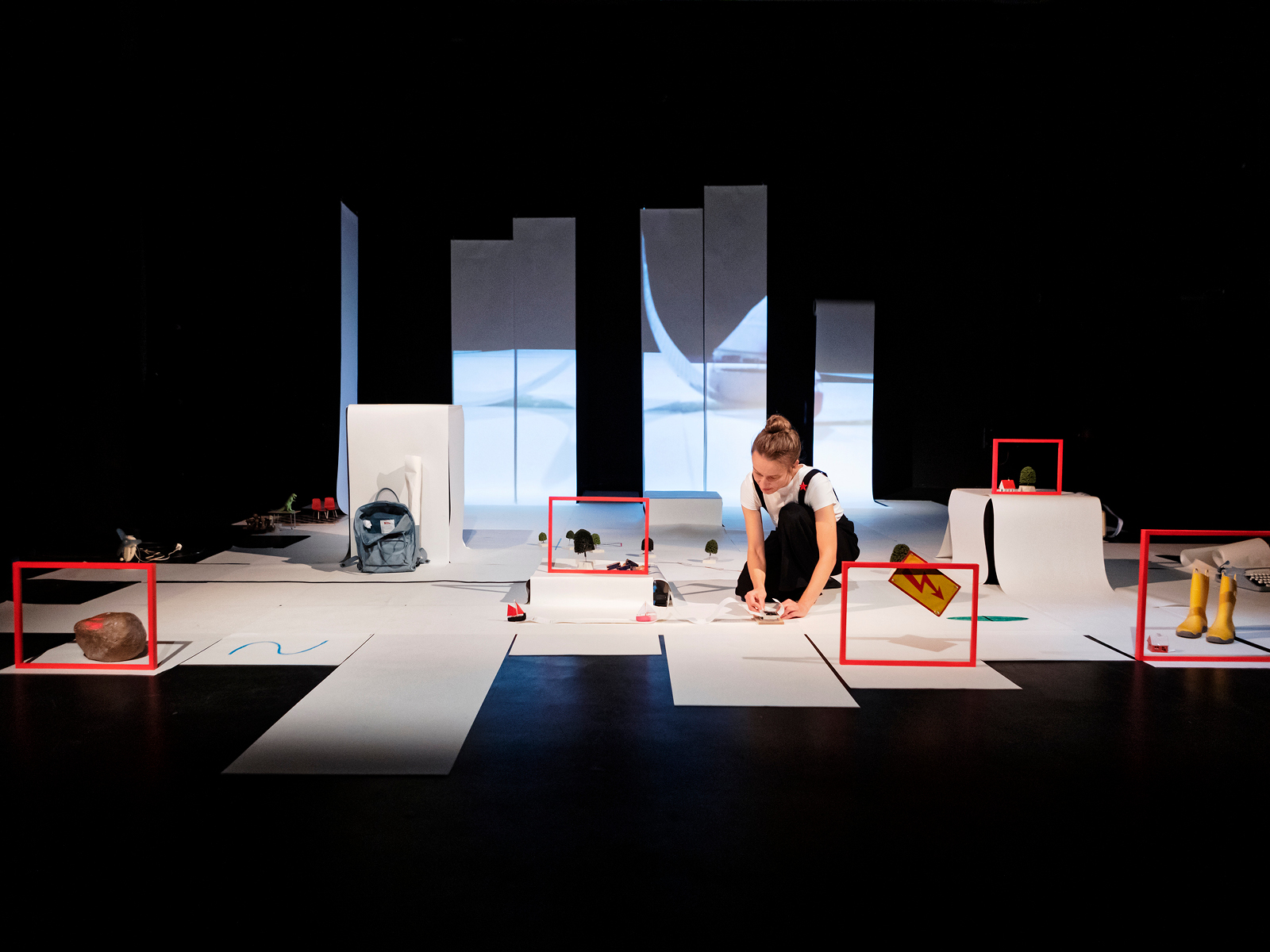Bühnenbild in der Gesamtansicht. Papierbahnen auf dem Boden und im Hintergrund an der Wand, wo sie als Projektionsfläche dienen. Die Spielerin kniet in der Mitte der Bühne, um sie herum sind kleine Gegenstände in rotem Holzrahmen abgebildet.
