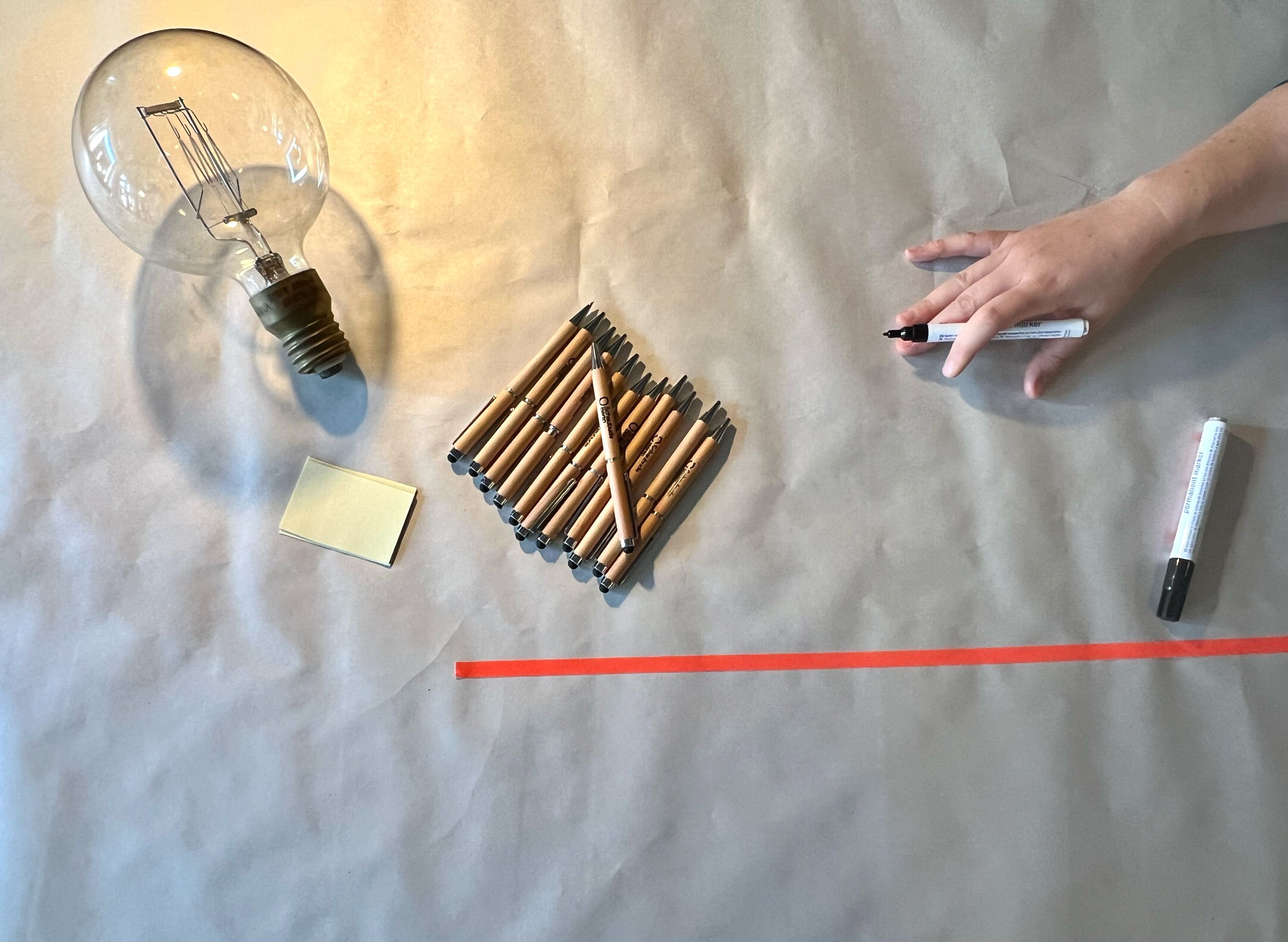 Ein Tisch mit einer Glühbirne, Stiften und einer Hand, die im Begriff ist etwas auf die Papiertischdecke zu schreiben.