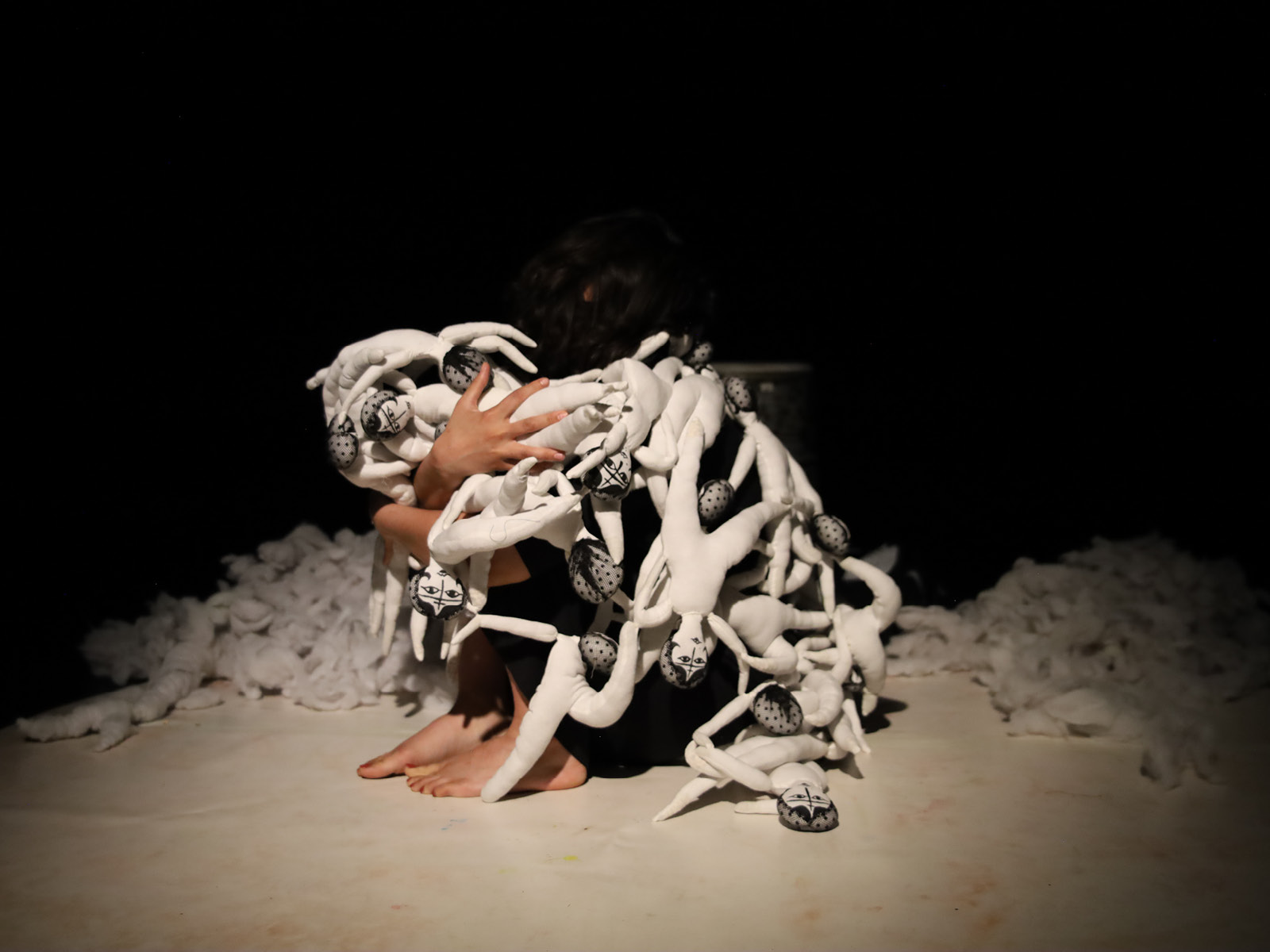 A person sits huddled on the floor. Her body is covered with small white dolls.