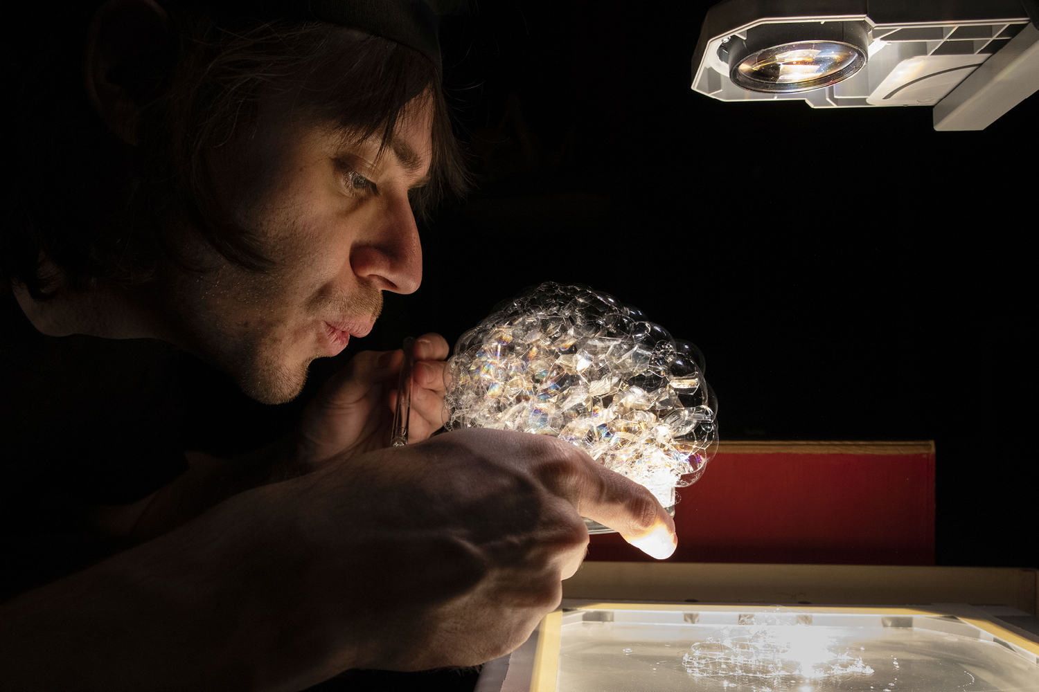 Ein Performer bläst mit einem Strohhalm Schaumblasen in einer Glasschale, die von einem Overhead-Projektor beleuchtet wird.