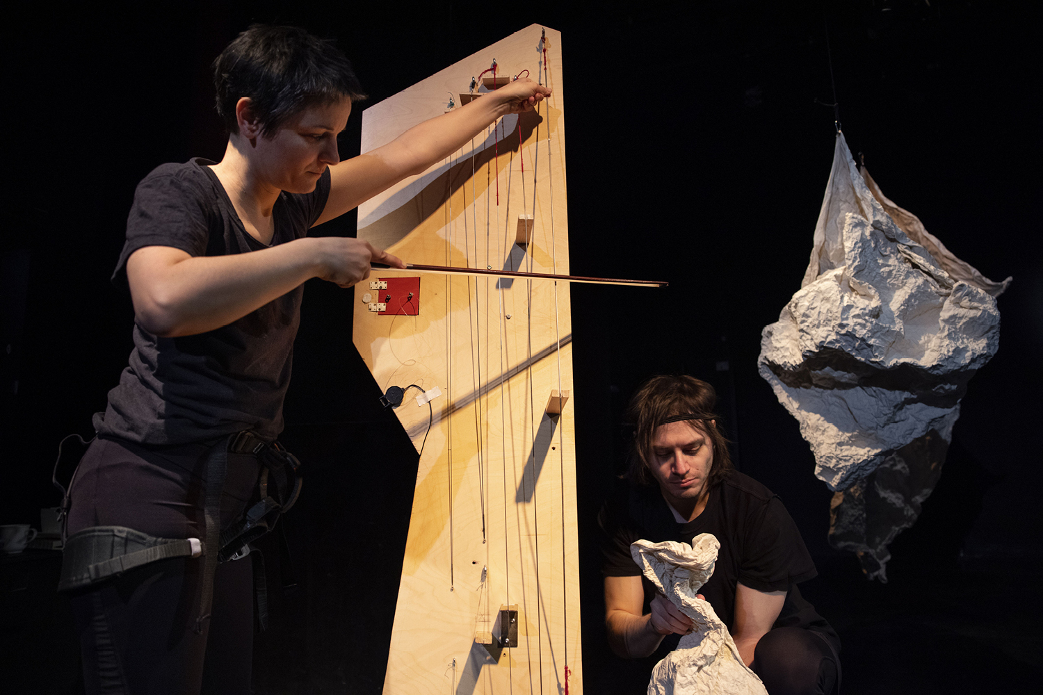 Performerin steht und streicht mit Geigenbogen die Saiten eines selbstgebauten Holzinstruments. Rechts daneben hockt ein weiterer Spieler, der mit den Händen ein papierartiges Material animiert als ob es eine Puppe wäre.