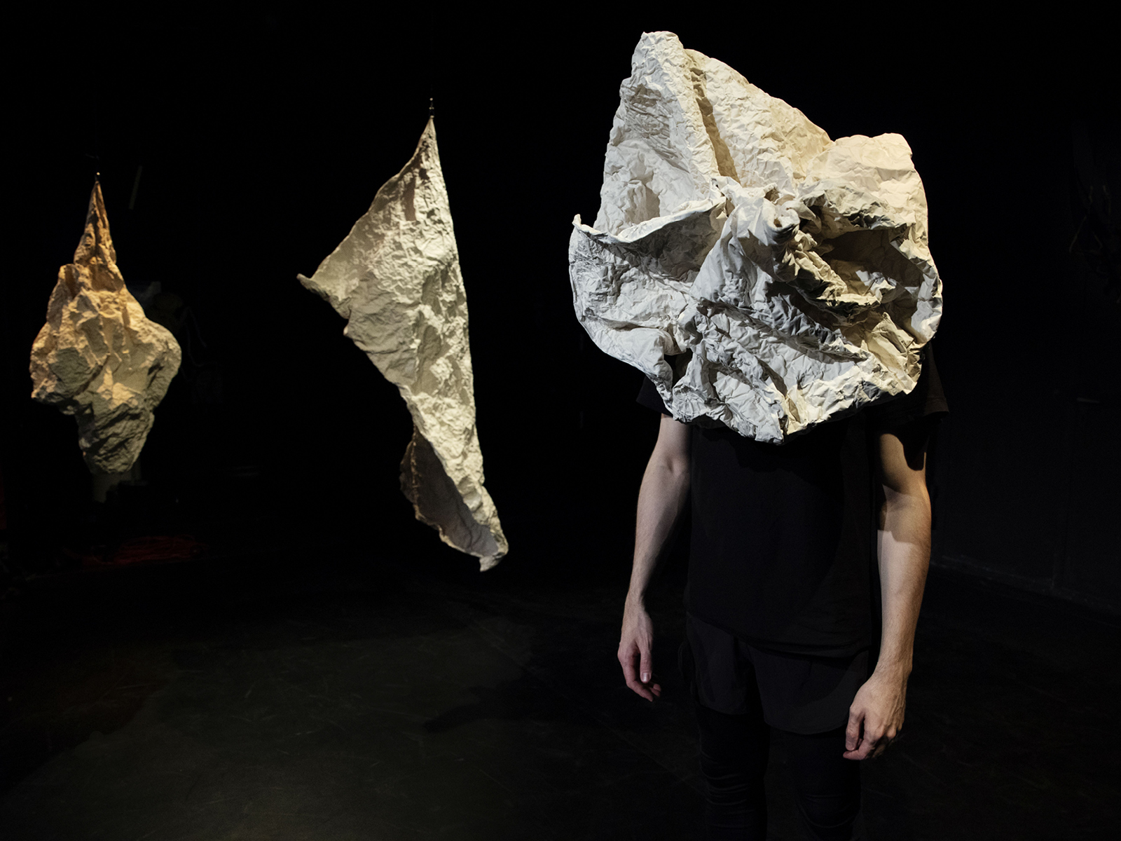 Performer mit papierähnlichem Material über dem Kopf; von ihm sind nur die Arme und schwarze Kleidung sichtbar. Im Hintergrund sind zwei von der Decke hängende weitere papierähnliche Formen zu sehen.